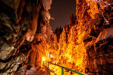 Breitach Gorge at night illuminated by warm lights, featuring rugged rocks and snow-covered trees. | © Kleinwalsertal Tourismus eGen • Breitachklamm • Photographer: Domink Berchtold