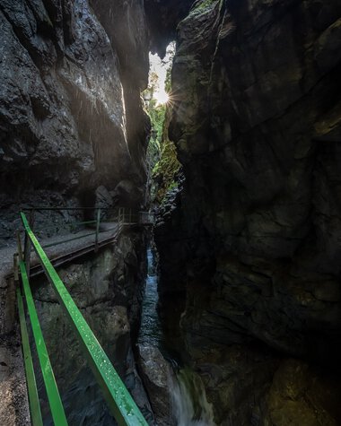 View through the Breitach Gorge in summer with waterfall and rugged rock walls, sun visible in the sky. | © Kleinwalsertal Tourismus eGen • Breitachklamm • Photographer: Dominik Berchtold
