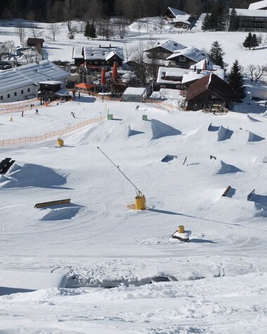 Snowpark at Crystal Ground with various jumps and obstacles in the snow, surrounded by a snowy mountain landscape and buildings. | © Kleinwalsertal Tourismus eGen • Crystal Ground
