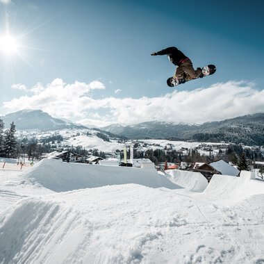 Snowboarder airborne over a jump ramp in a snowy winter landscape under sunlight. | © Kleinwalsertal Tourismus eGen • Crystal Ground • Photographer: Stefan Eigner