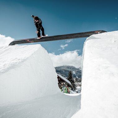 Skateboarder jumping over a snow gap with snow walls on both sides, under clear sky. The scene is set on a winter ski slope. | © Kleinwalsertal Tourismus eGen • Crystal Ground • Photographer: Stefan Eigner