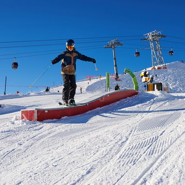 Skier skier riding on a snowboard feature at a ski resort in Oberstdorf Kleinwalsertal, with ski lifts and slopes in the background. | © Kleinwalsertal Tourismus eGen • Oberstdorf Kleinwalsertal Bergbahnen