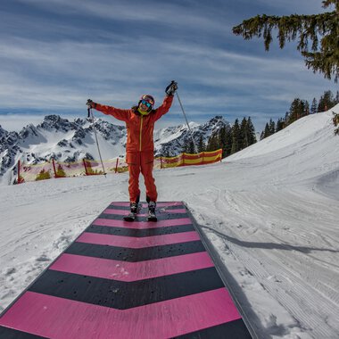 Skier person in red on a snow-covered slope in Oberstdorf, with mountains in the background and a blue sky. | © Kleinwalsertal Tourismus eGen • Oberstdorf Kleinwalsertal Bergbahnen
