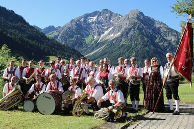 Musikkapelle in traditioneller Tracht vor einer Bergkulisse mit hohen Bergen und Wäldern, Gruppe mit Instrumenten und Banner. | © Kleinwalsertal Tourismus eGen • Musikverein d'Hirschegger