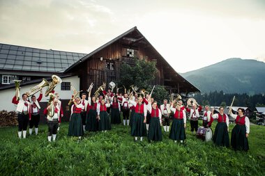 Trachtenkapelle Riezlern in traditioneller Kleidung spielt auf einer grünen Wiese vor einem Bauernhaus mit Bergen im Hintergrund. | © Kleinwalsertal Tourismus eGen • Fotograf: Oliver Farys