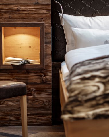 Innenansicht eines gemütlichen Zimmers mit einem Bett, Kopfteil in Schwarz, und einer Holzbox an der Wand, die ein Buch enthält. | © Bergchalet