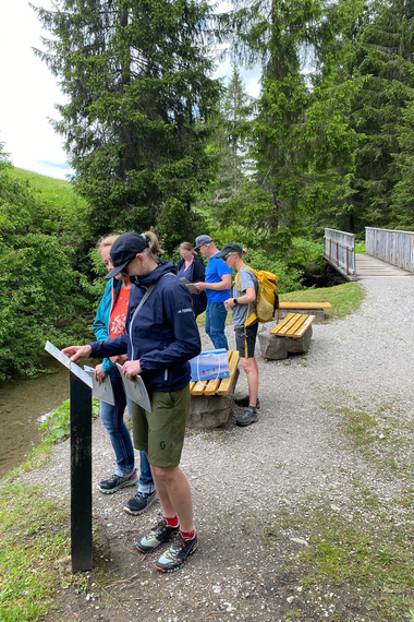 Gruppe von Menschen liest Informationen an einer Infotafel in einer grünen Waldlandschaft beim Wandern. | © Kleinwalsertal Tourismus eGen