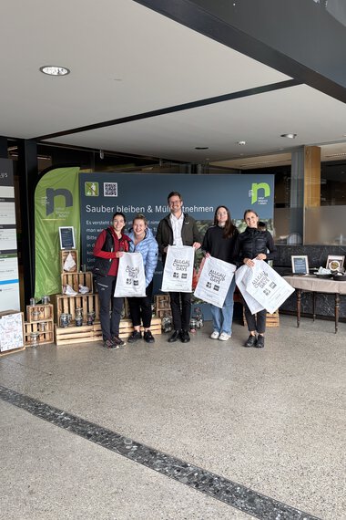 Vier Personen bei einer Umweltaktion im Eingangsbereich eines Gebäudes, halten Tüten mit Recycling-Material. Hinter ihnen eine blaue Werbetafel. | © Kleinwalsertal Tourismus eGen