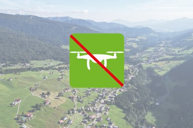 Verbotsschild für den Betrieb von Drohnen, grün mit rotem Schrägstrich, im Hintergrund eine Berg- und Tallandschaft. | © Kleinwalsertal Tourismus eGen