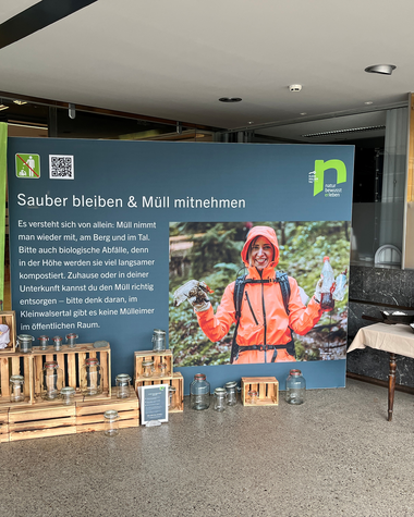 Informationsstand zur Müllkampagne, mit einem großen blau-grünen Poster, Holzboxen, und zwei Tischen mit Tablets, im Innenraum. | © Kleinwalsertal Tourismus eGen