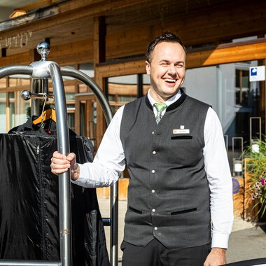 Hotelmitarbeiter am Rezeptionbereich vor einem modernen Holzgebäude, lachend mit einem Gepäckwagen. | © Kleinwalsertal Tourismus eGen • Fotograf: Werner Krug