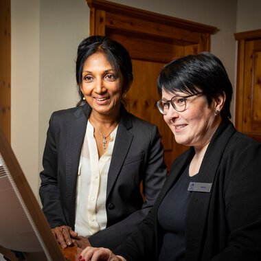 Zwei Frauen im Büro, eine arbeitet am Computer, beide lachen und wirken freundlich. Die Umgebung ist warm und einladend. | © Kleinwalsertal Tourismus eGen • Fotograf: Werner Krug