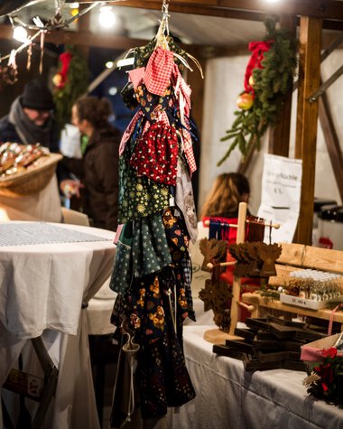 Christmas market in Mittelberg with festively decorated stalls, colorful fabrics, Christmas decorations, and visitors during Advent. | © Kleinwalsertal Tourismus eGen • Photographer: Dominik Berchtold