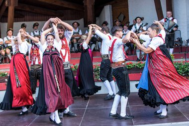 Tanzgruppe in traditionellen Trachten beim Alphornfestival 2018, tanzend auf einer Bühne, umgeben von Musikern. | © Kleinwalsertal Tourismus eGen • Fotograf: Frank Drechsel