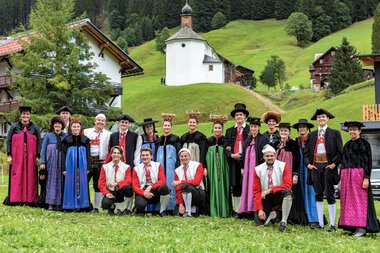 Gruppe von Menschen in traditioneller Tracht vor malerischer Bergkulisse mit Kirche und Häuser im Hintergrund, wahrscheinlich bei einem Alphornfestival 2022. | © Kleinwalsertal Tourismus eGen • Fotograf: Frank Drechsel