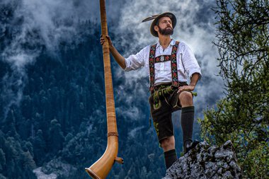 Mann in traditioneller Tracht mit Alphorn auf einem Felsen im Gebirge, Blick nach oben, im Hintergrund Wolken und Bäume. | © Loisach Marci • Anton Ostler