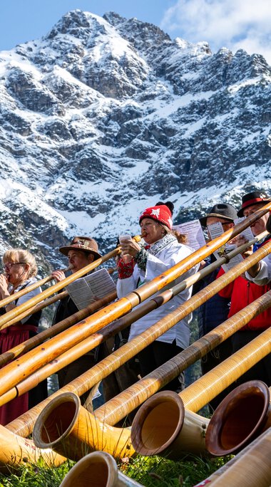 Menschen beim Alphornfestival in den Bergen, mit Berggipfel im Hintergrund, spielen auf traditionellen Alphorninstrumenten. | © Kleinwalsertal Tourismus eGen • Fotograf: Frank Drechsel